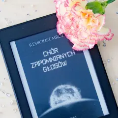 Elektroniczna książka z tytułem "Chór zapomnianych głosów" Remigiusza Mroza, ozdobiona kwiatem i rozsypanymi kryształkami.