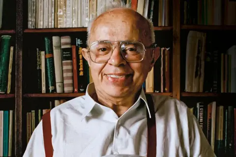 Uśmiechnięty Stanisław Lem, autor powieści Solaris, siedzi przed regałami pełnymi książek.