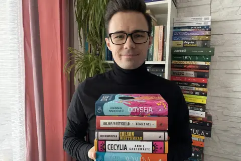 Mężczyzna w okularach trzyma stos książek, wśród nich "Odyseja" i dzieła Colsona Whiteheada. W tle regały pełne literatury, przypominające o bogactwie polskiej i światowej prozy, jak u Bolesława Prusa.