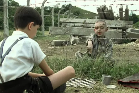 Chłopiec w pasiastej koszuli wskazuje na planszę do gry. Obok siedzi inny chłopiec w białej koszuli i szelkach.