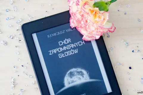 Elektroniczna książka z tytułem "Chór zapomnianych głosów" Remigiusza Mroza, ozdobiona kwiatem i rozsypanymi kryształkami.