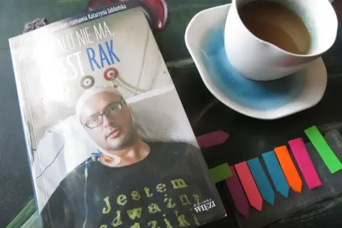 Książka "Żalu nie ma, jest rak" z wizerunkiem pacjenta, obok filiżanka kawy i kolorowe zakładki.