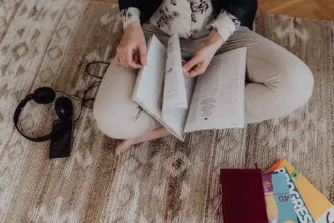 Kobieta czyta książkę, obok leżą słuchawki i telefon. W tle widać kolorowe książki, idealne do zanurzenia się w literaturze francuskiej klasyce.