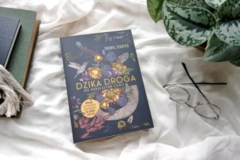 Okładka książki "Dzika droga" z motywami roślinnymi i zwierzęcymi, obok okulary i inne książki.