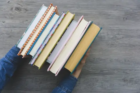 Ręce w dżinsowej kurtce trzymają stos kolorowych książek. To idealne nowości warte przeczytania na jesienne wieczory.