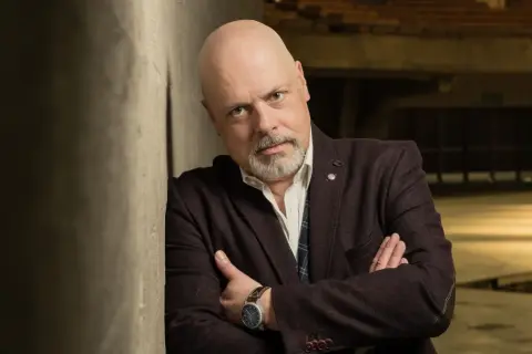 Marek Krajewski w eleganckim stroju, oparty o ścianę, zamyślony. W tle industrialne wnętrze, sugerujące jego życie prywatne.