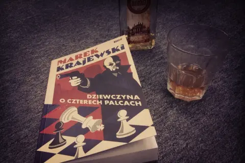 Powieść Marka Krajewskiego "Dziewczyna o czterech palcach" leży obok szklanki z bursztynowym trunkiem.